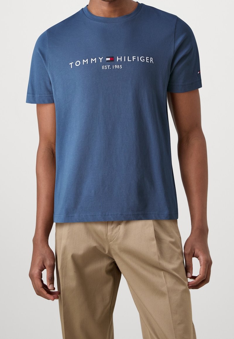 Blue cotton t-shirt with short sleeves featuring "TOMMY HILFIGER EST. 1985" logo in white and red, paired with beige trousers.