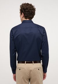 Marineblaues Langarmhemd mit klassischem Kragen, glatter Textur und knöpfbaren Manschetten, kombiniert mit khakifarbenen Hosen mit Gesäßtaschen.