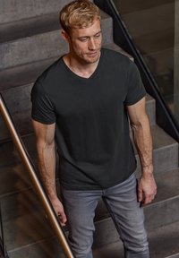 Zwarte v-hals t-shirt van een zachte stof, gecombineerd met lichtgrijze jeans. De setting bevat betonnen trappen en een metalen leuning.