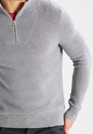 Homme portant un pull en maille texturée gris clair avec col zippé au quart et poignets côtelés, associé à un pantalon sombre, sur fond uni.