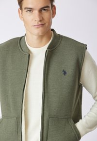 Gilet senza maniche verde oliva con zip frontale e due tasche, sovrapposto a una camicia a maniche lunghe color crema a coste. Presenta un logo ricamato.