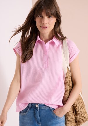 Femme vêtue d'une chemise rose à manches courtes boutonnée, un jean bleu et portant un sac à bandoulière marron tressé, debout devant un fond uni.