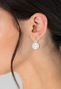 Guldörhängen med en vit pärla som hänger ner, med en slät yta och rundad design, visade på en models öra.