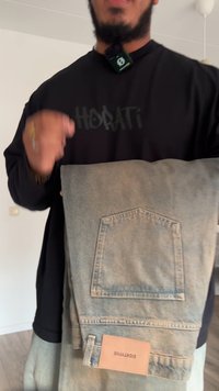 Világoskék farmer vászontáska, melynek kiemelkedő zsebe, bőrből készült címkéje és puha tapintása van. Fekete, laza fazonú ing viselve.