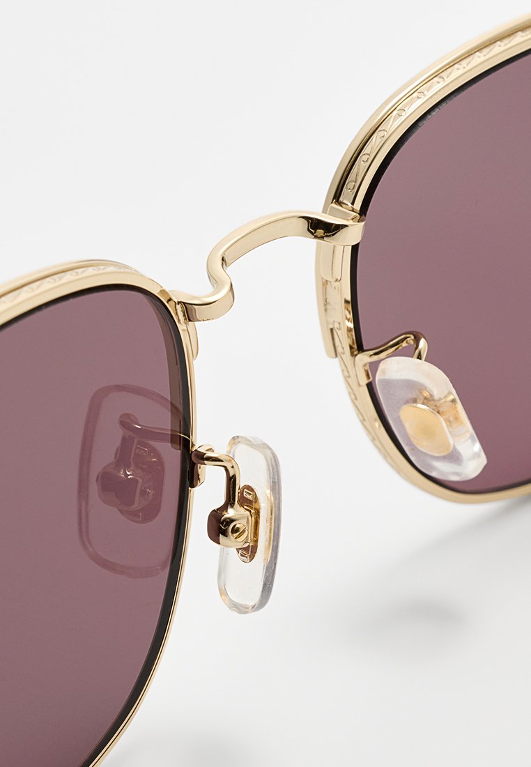 Goldfarbene Sonnenbrille mit dunkellila Gläsern. Zu den Merkmalen gehören eine doppelte Brücke und verstellbare klare Nasenpolster für zusätzlichen Komfort.