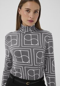 Grå rullekravetrøje med hvide geometriske mønstre, med lange ærmer og en tæt pasform, kombineret med et sort bælte i taljen.