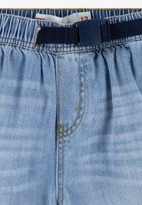 Pantalones de mezclilla azul claro con cinturilla elástica y cinturón ajustable en color marino. Presentan costuras amarillas y un pequeño detalle cosido cerca de la cremallera.