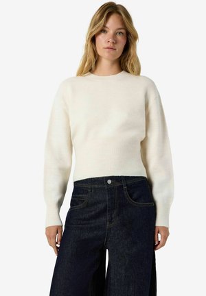 Femme portant un pull en tricot crème à manches longues et un jean large taille haute bleu foncé, debout devant un fond uni.