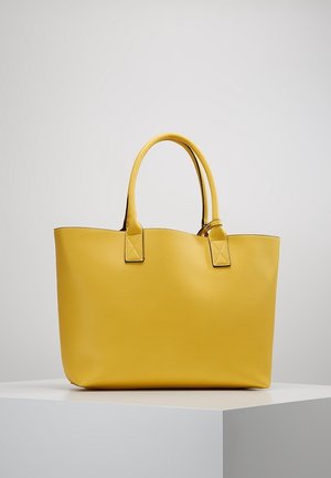 Bolso tote de piel amarillo con asas dobles, mostrado sobre un pedestal blanco contra un fondo gris claro liso.