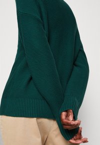 Pull vert foncé en tricot avec motif texturé, poignets et bas côtelés. Mains visibles derrière, portant un pantalon beige.
