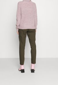 Gebreide roze trui met een hoge kraag, bruine broeken en glanzende roze enkellaarsjes met blokhakken. Simpele, gelaagde texturen en kleuren.