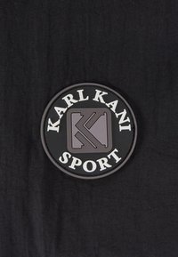 Svart cirkulär patch med "KARL KANI" högst upp, "SPORT" längst ner och en stiliserad logotyp i mitten, med vita och grå accenter.
