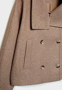 Manteau en laine marron avec col large, boutons croisés en écaille de tortue et une poche latérale rectangulaire sur le devant gauche.