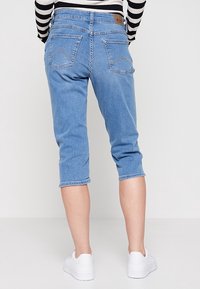 Pantalones capri de mezclilla azul claro con corte recto, dos bolsillos traseros con un patrón de zigzag y dobladillo enrollado. Se llevan con zapatillas blancas.