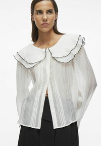 Camisa blanca de botones con un gran cuello volante con ribete negro, tejido texturizado con rayas verticales y mangas largas.