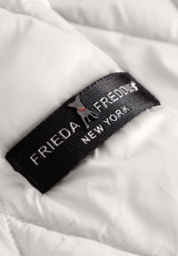 Witte pluche stof met een zwart en zilveren label met de tekst "FRIEDA FREDDY NEW YORK," met een hondenafbeelding in het logo.