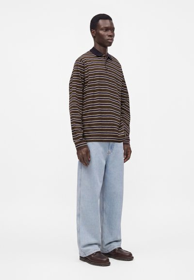 Homme debout portant une chemise à rayures marron à manches longues, un jean large bleu clair et des chaussures marron foncé, sur un fond blanc uni.