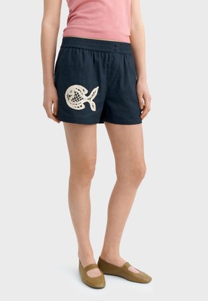 Žena oblečená v námorníckych modrých šortkách s bielym dizajnom ryby na ľavej nohe, ružovom tielku bez rukávov a hnedých plochých topánkach, stojaca pred jednoduchým pozadím.