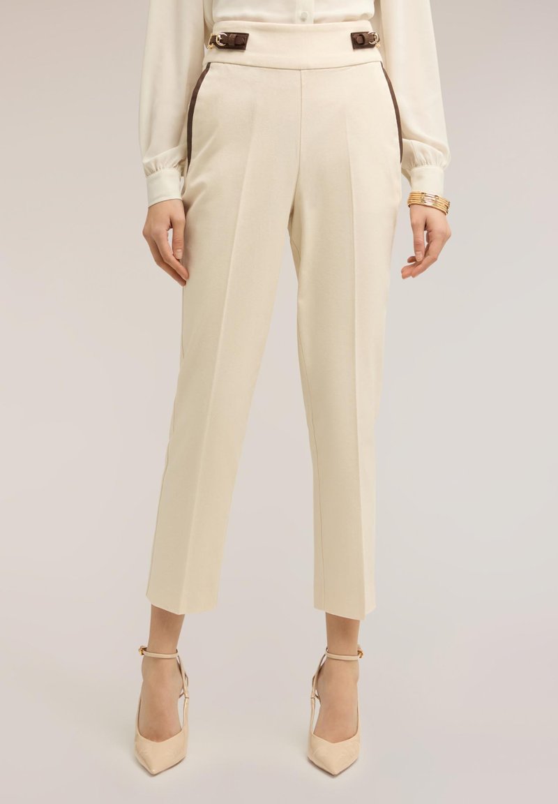 Woman wearing high-waisted cream trousers with brown buckle details, paired with cream pointed-toe ankle-strap heels and a cream blouse.