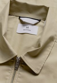 Jachetă bej cu închidere prin fermoar, guler plat și un eticheta pe care scrie "Matiniqué". Materialul are o textură netedă și o strălucire subtilă.