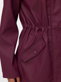 Burgundy vattentät jacka med insydd midja, långa ärmar och en tryckknappsficka på sidan. Slät, glänsande yta.