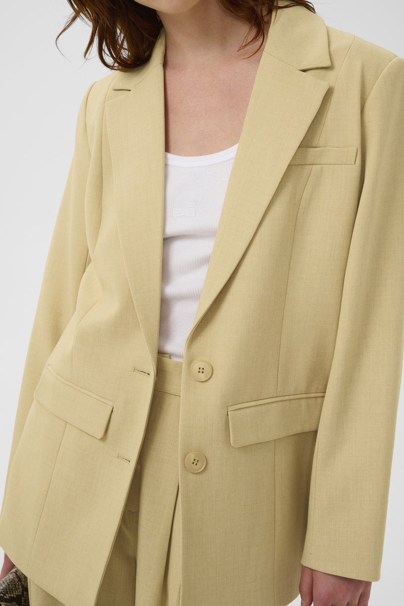 Femme portant un blazer beige clair cintré avec deux boutons et des poches à rabat, par-dessus un débardeur blanc côtelé.