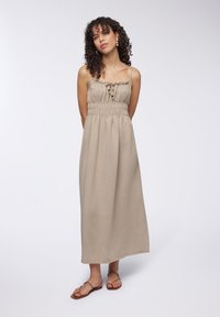 Abito maxi beige in tessuto leggero con vita smock, spalline regolabili e dettaglio dei bottoni frontali. Modello che indossa sandali flat.