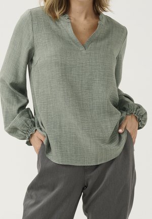 Vrouw draagt een loszittende lichtgroene blouse met lange mouwen en geplooide manchetten, en donkergrijze broek, handen in de zakken, neutrale achtergrond.