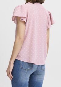 Różowa bluzka z krótkim rękawem z fakturowanym wzorem w groszki, o luźnym kroju i falbanianych rękawach. Noszona z niebieskimi dżinsami.