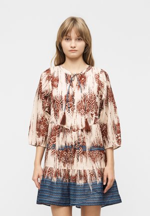 Młoda kobieta w sukience z długim rękawem o brązowo-kremowym geometrycznym wzorze i niebieskim paskowanym obszyciu na dole, stojąca na jednolitym tle.