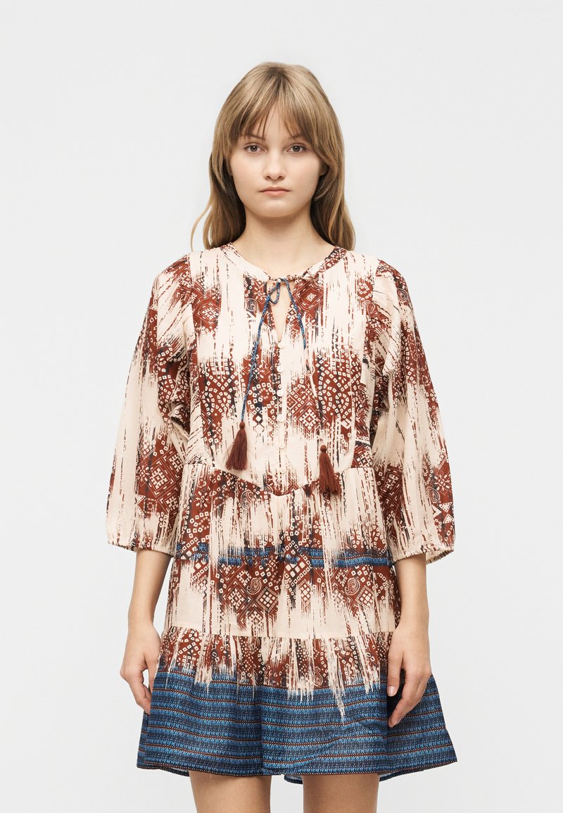 Νεαρή γυναίκα που φοράει φόρεμα μακρυμάνικο με καφέ και κρεμ γεωμετρικό σχέδιο και μπλε ρίγες στο τελείωμα, στέκεται μπροστά σε απλό φόντο.