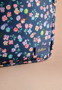 Tengerészkék szövet táska virágmintával, rózsaszín, lila és zöld díszítésekkel; sötét címkével, amelyen a „Cath Kidston” domború felirat található.