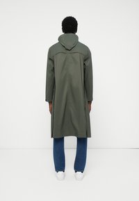 Lungo cappotto verde oliva con cappuccio, tessuto morbido, spacco laterale, indossato sopra jeans blu e sneakers bianche, visto da dietro.