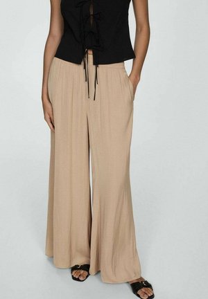 Mujer con pantalones anchos beige, top negro sin mangas con lazos delanteros y sandalias negras con los dedos descubiertos, de pie con una mano en el bolsillo.