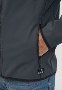 Veste softshell gris foncé avec fermeture éclair à l'avant, poches latérales et ourlet noir. Présente un petit patch logo près de la fermeture éclair.