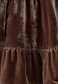 Brunt fløjlstof med en blød, skinnende tekstur, der har en tiered flæse ved kanten. Materialet fremstår fyldigt og luksuriøst.