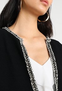 Czarny kardigan z teksturowanej tkaniny, ozdobiony dekoracyjną białą i czarną lamówką przy dekolcie. Nosi się go na białą bluzkę.