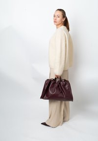 Burgundy leather handbag with a gathered top, held by a person in a cream sweater and beige trousers against a white background.