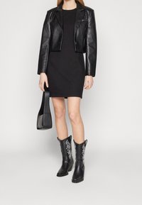 Czarna skórzana kurtka krótka noszona na czarnej dopasowanej sukience, w zestawieniu z czarnymi kowbojkami z białym haftem i czarną torbą na ramię.