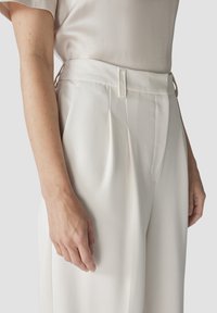 Weiße, gefaltete Hose mit einer glatten, weichen Textur und einem hohen Bund, ausgestattet mit seitlichen Haken zum Verschließen und minimaler Detailgestaltung.