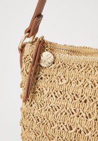 Ljusbrun vävd halmhandväska med en texturerad yta, guldfärgad dragkedja och ett läderhandtag. Har en dekorativ guldkraft som är fäst.