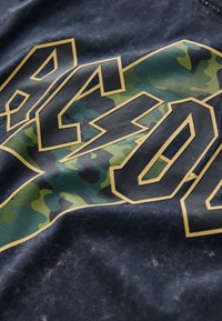 Marineblauwe t-shirt met vet gele tekst en camouflagepatronen in groen en donkere tinten, tegen een gestructureerde achtergrond.
