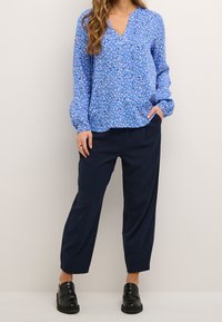 Blå mønstret bluse i let stof med V-udskæring, lange ærmer og elastiske manchetter, parret med marineblå bukser og sorte loafers.