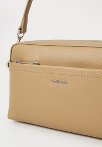 Sac à main en faux cuir beige avec une texture lisse, de forme rectangulaire, une poche avant zippée et un logo Calvin Klein en argent.