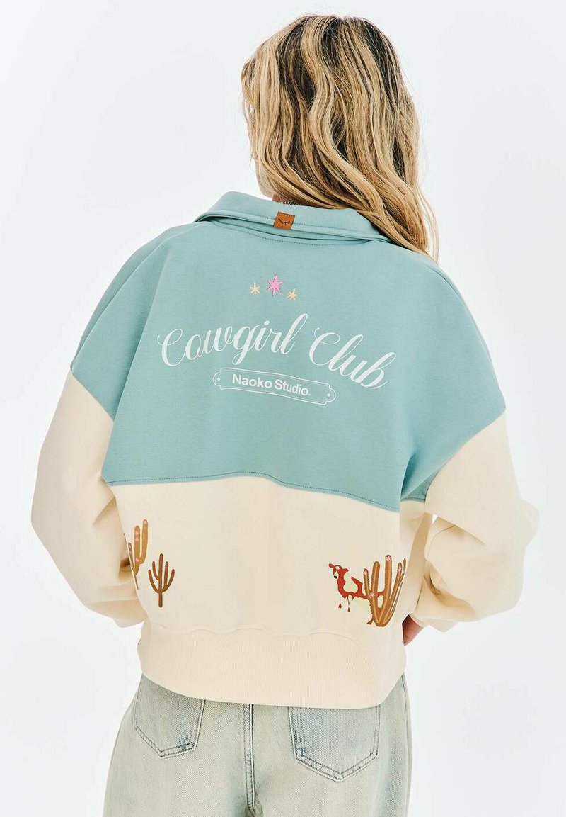 Žena s dlouhými blond vlasy nosí dvoubarevnou mikinu s nápisem "Cowgirl Club" a vzory kaktusu a koně na zádech.