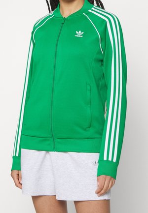 Personne portant une veste de sport Adidas verte avec des bandes blanches sur les manches et une jupe gris clair, debout devant un fond uni.