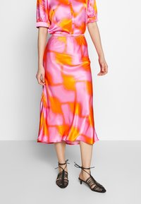 Jupe midi en satin avec un motif abstrait rose et orange vif, associée à des sandales noires à lanières pour un look moderne.