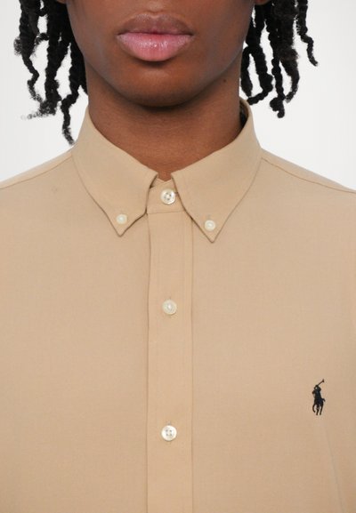 Camisa bege de botão com colarinho pontudo, apresentando um pequeno logotipo preto. Botões claros e textura suave são visíveis.