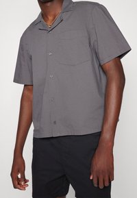 Krátký rukáv, šedá košile na knoflíky vyrobená z tkané látky. Obsahuje jednu náprsní kapsu a volný střih. Spárováno s tmavými šortkami.