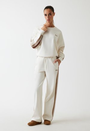 Cremiges Sweatshirt- und Hosen-Set mit braunen Seitenstreifen und Logo-Stickerei. Weicher Stoff, lässige Passform und seitliche Taschen an der Hose.